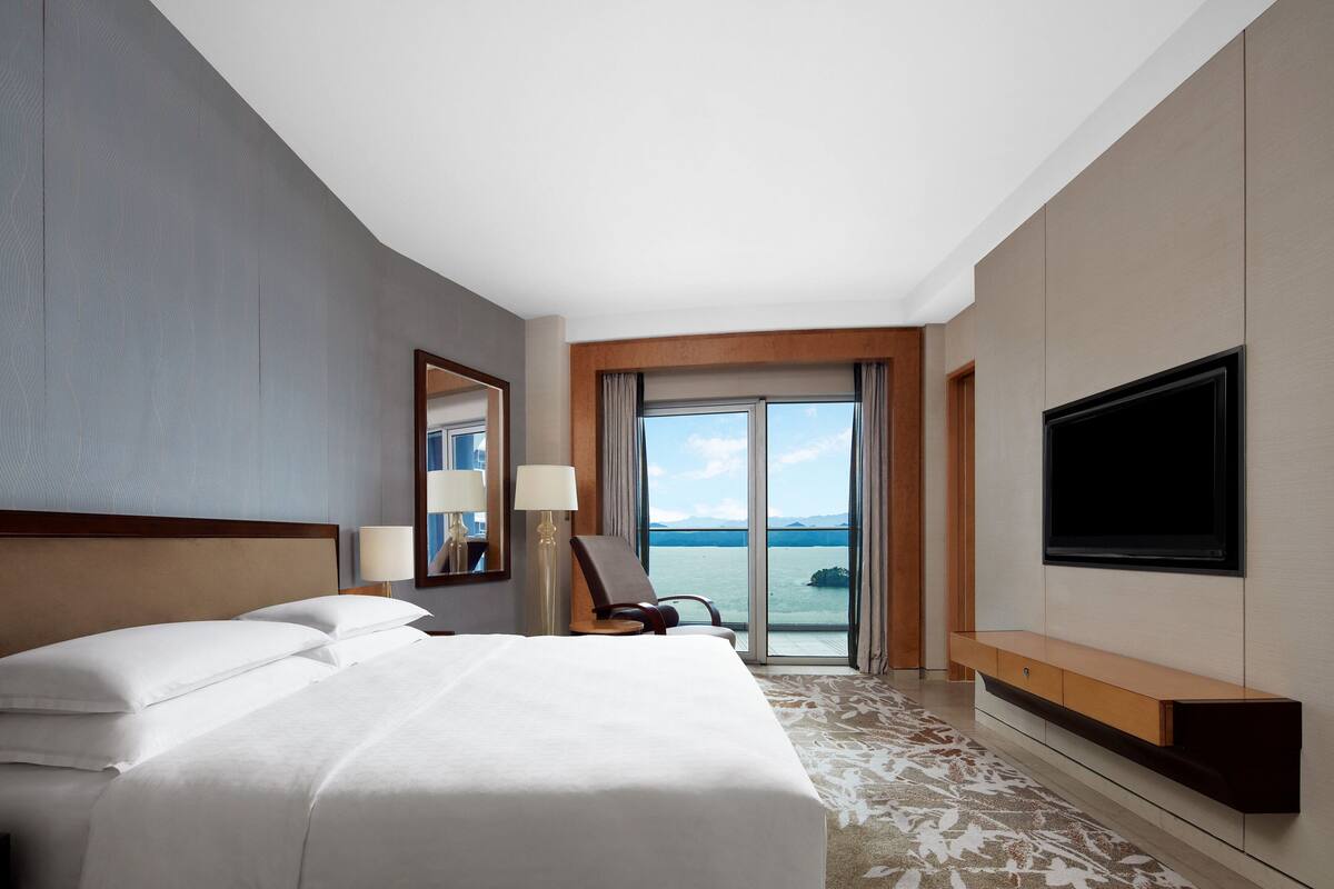 A hotel room with a large bed, a TV mounted on the wall, and a view of the sea through the window.