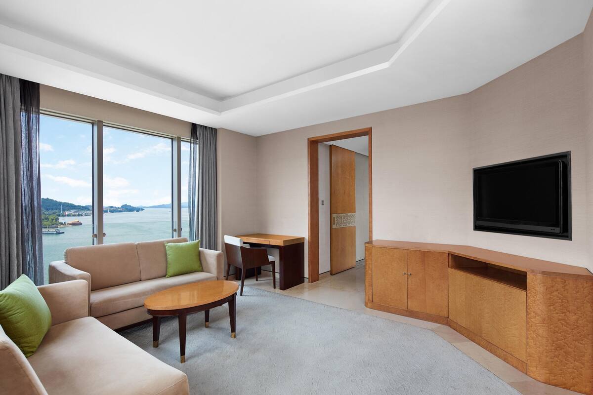 A modern hotel room with a large window offering a view of the sea, a sofa, a dining table, and a flat-screen TV mounted on the wall.