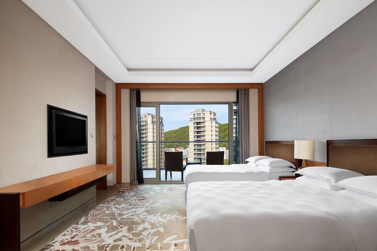 A hotel room with two beds, a TV, a desk, and a view of high-rise buildings.