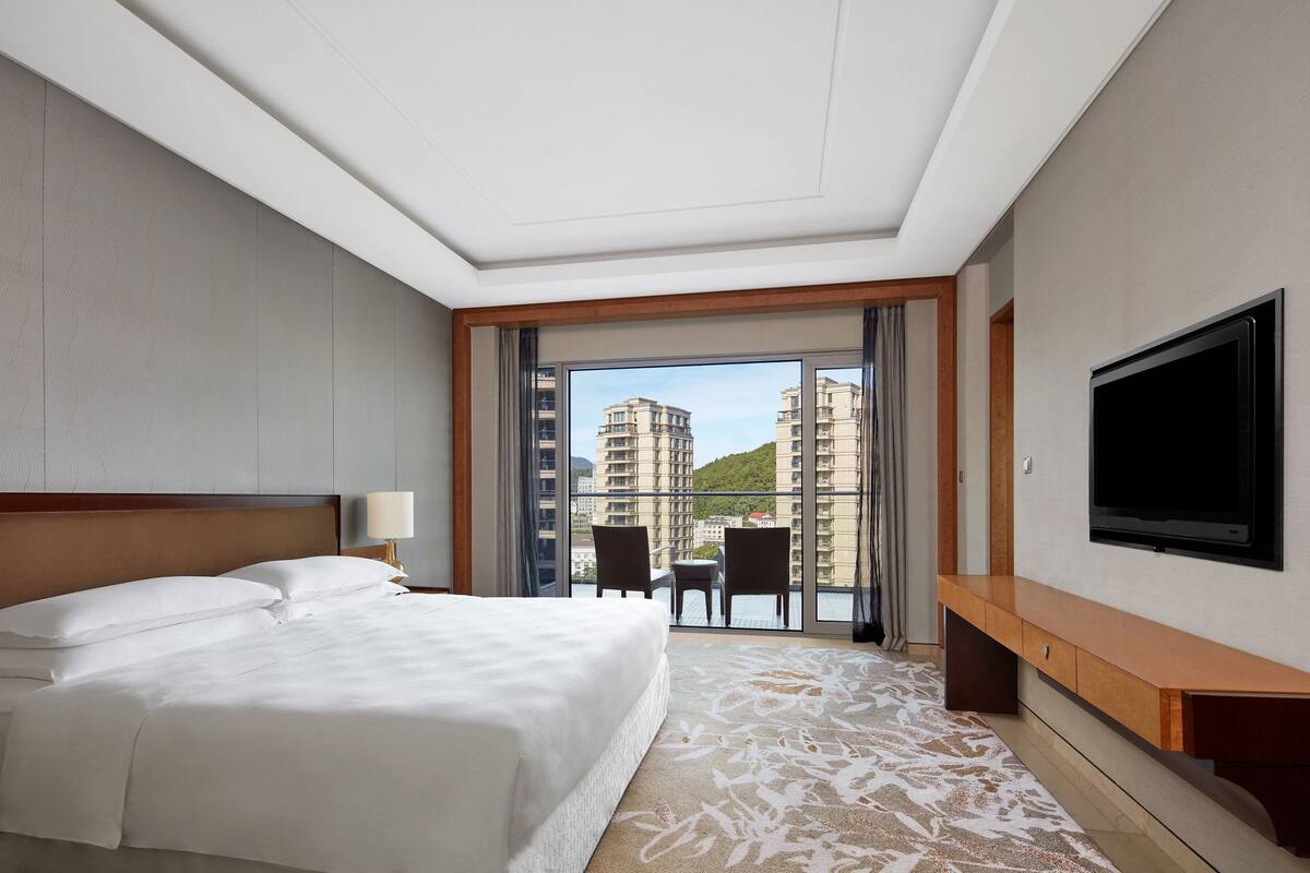 A hotel room with a large bed, a TV, a balcony with a view of buildings, and a desk.