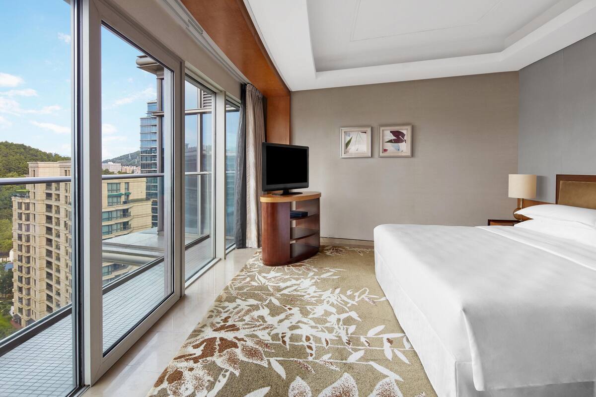 A hotel room with a large window, a bed, a TV, and a small table.