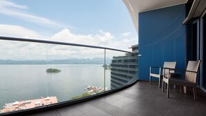 Un balcón con barandilla de vidrio curvo, dos sillas de mimbre y vistas a un lago con una isla distante y montañas.