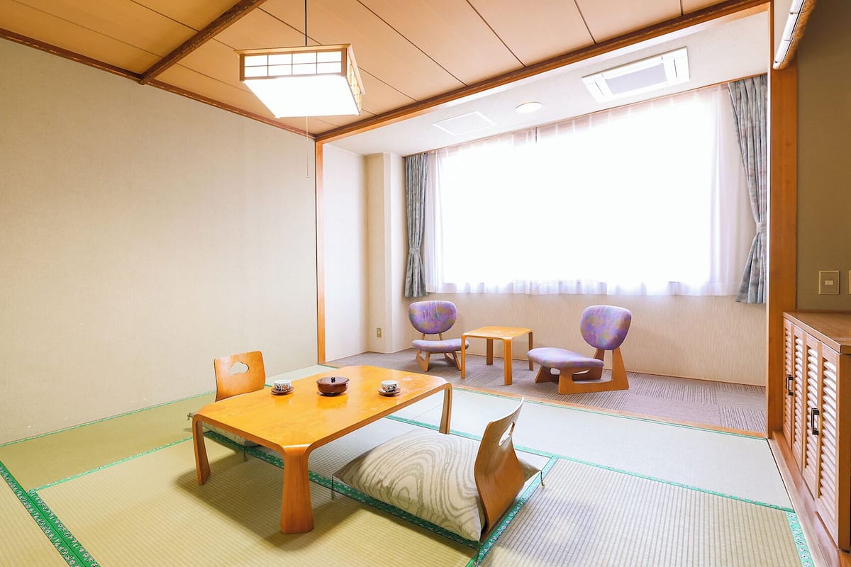 Une pièce de style japonais traditionnel, avec un sol recouvert de tatamis, une table en bois sur laquelle est disposé un service à thé, et deux chaises violettes.