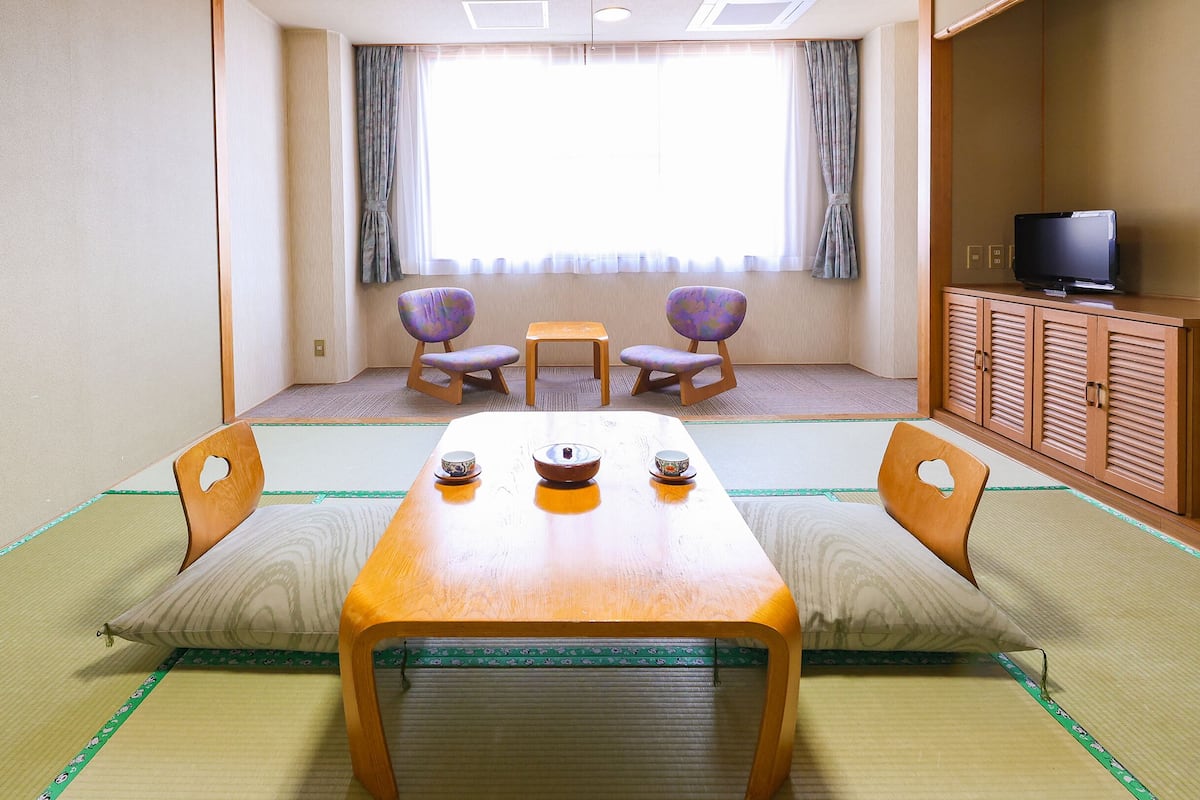 A traditional Japanese room with tatami flooring, a low table with cushions, two chairs, a small table, and a television on a cabinet.