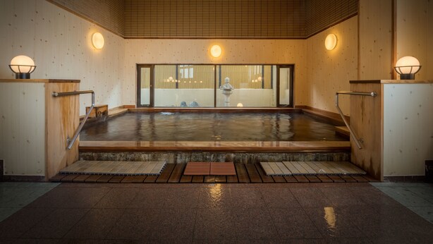 Un onsen japonés tradicional con paredes de madera, una fuente termal de piedra y piso de baldosas.