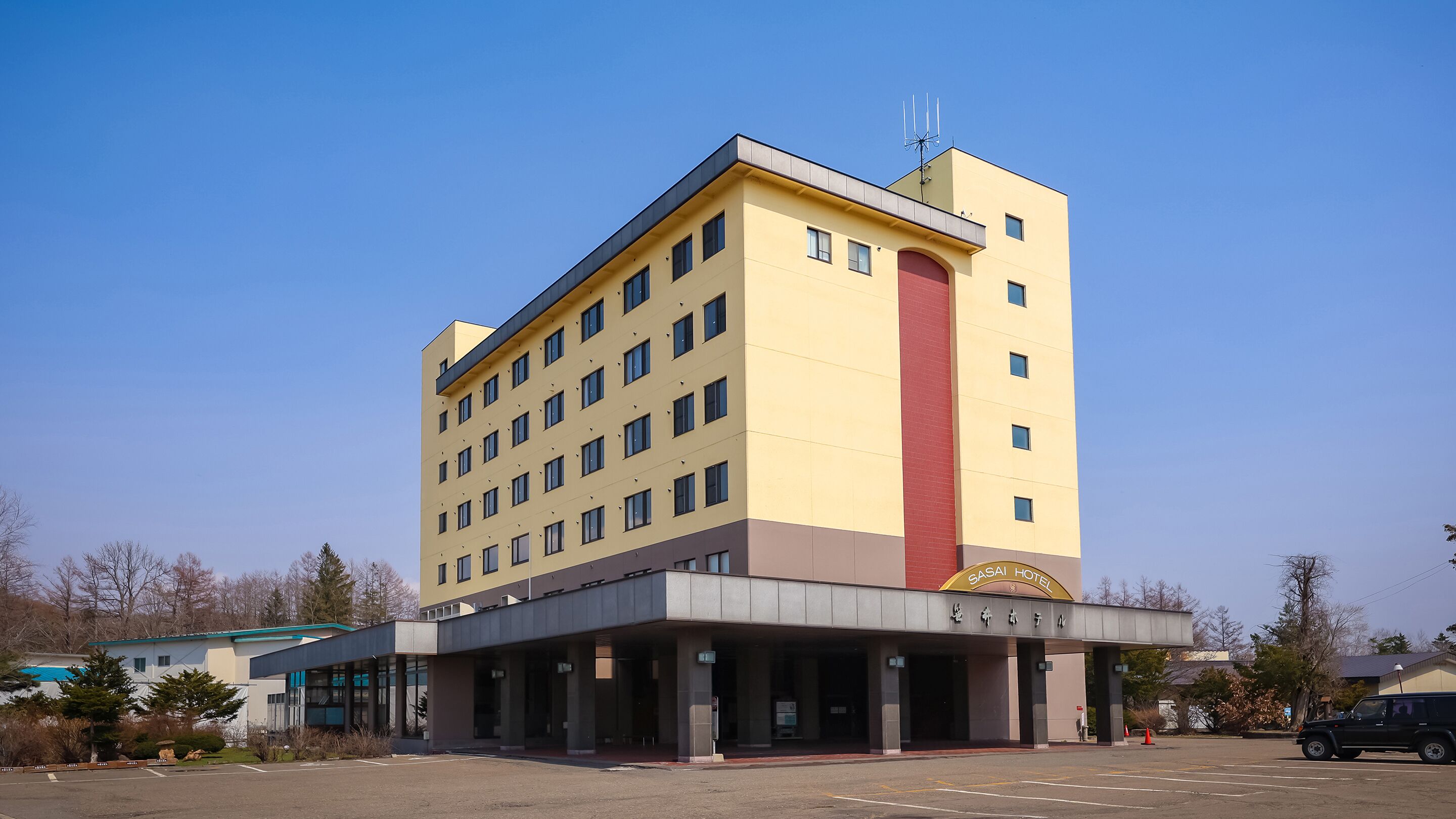 Ein mehrstöckiges Hotel mit flachem Dach, einem auffälligen roten vertikalen Abschnitt und einem Schild, das anzeigt, dass es sich um ein SASAI HOTEL handelt.