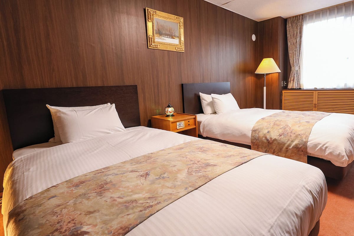 Une chambre d’hôtel avec deux lits, une table de chevet, une lampe, un tableau encadré et une fenêtre avec des rideaux.