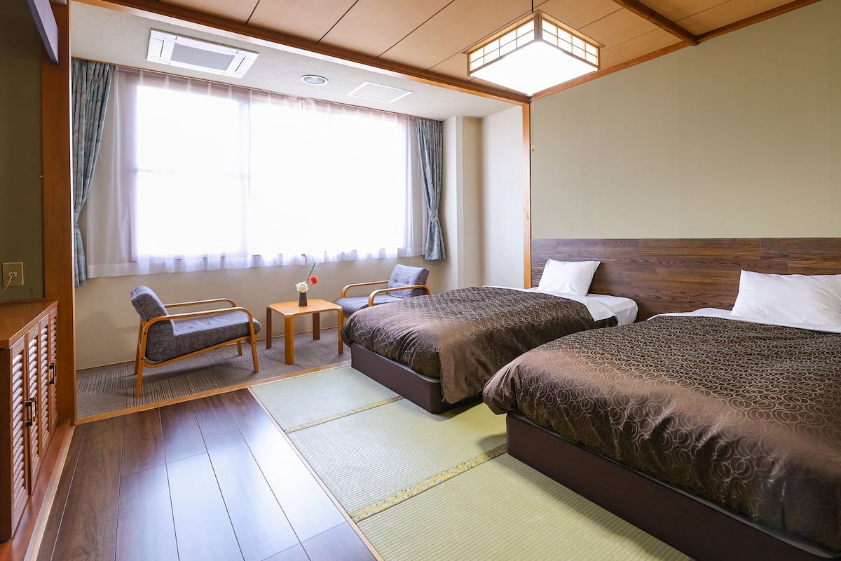 A hotel room with two beds, a small table with a vase, a chair, and a window with curtains.