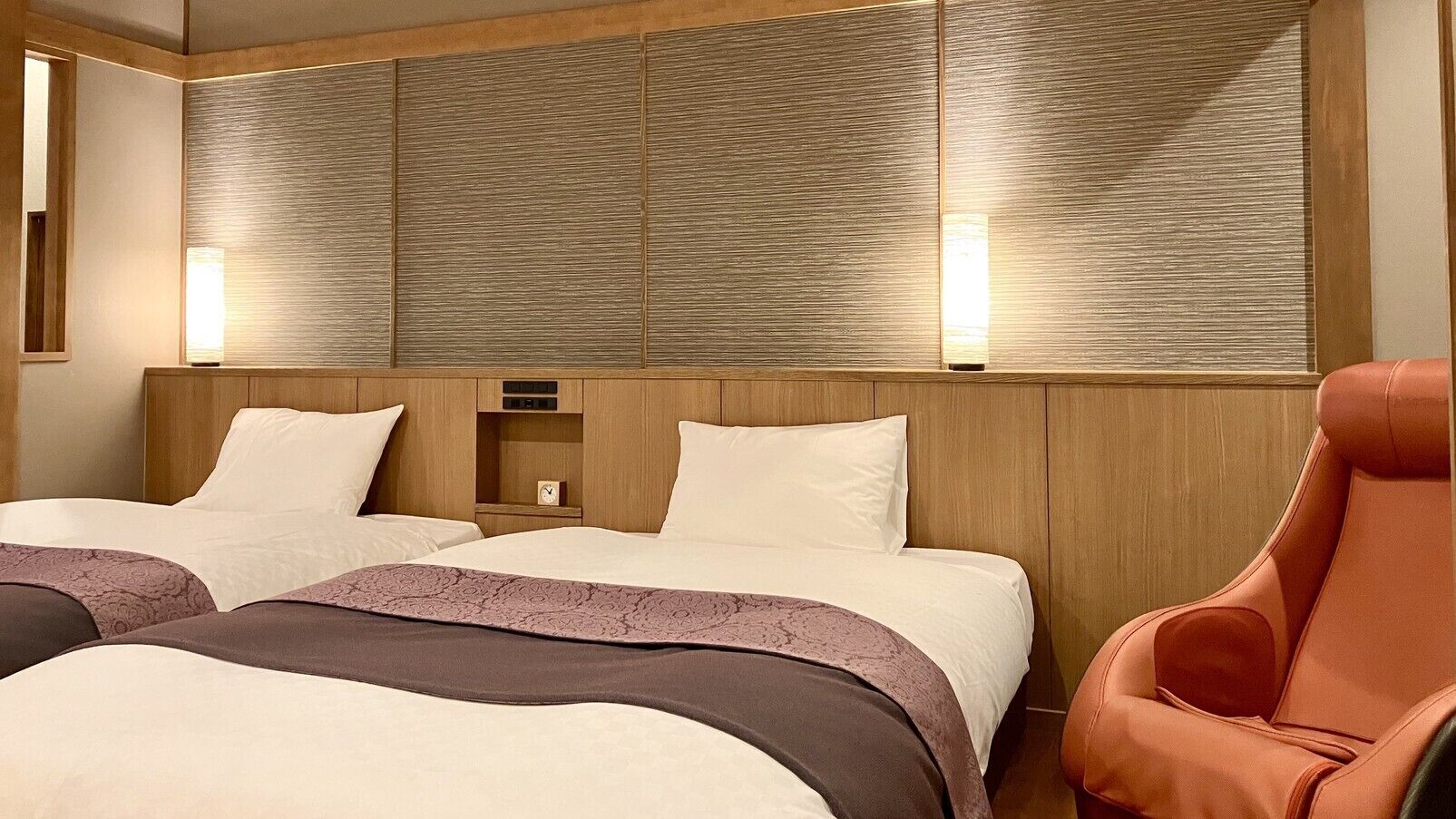 A hotel room with two beds, a wooden headboard, a nightstand, and a red armchair.