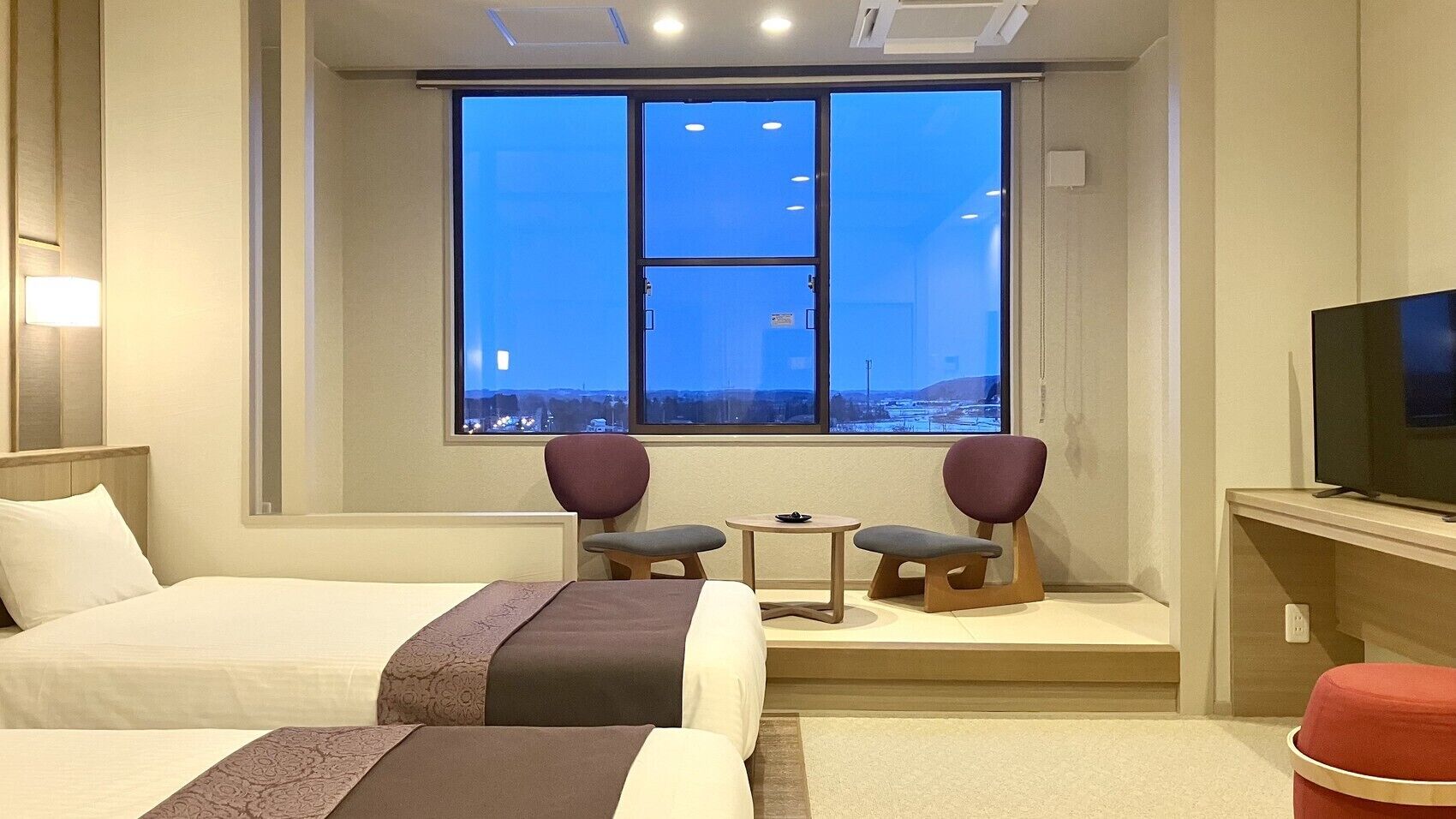 A hotel room with two beds, a TV, a window with a city view, and a small table with chairs.