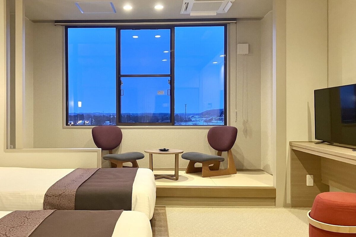 A hotel room with two beds, a TV, a window with a city view, and a small table with chairs.