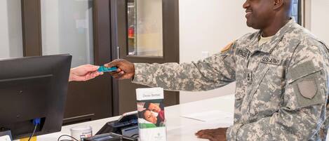 A person in military uniform at a counter with a computer monitor, a card reader, and a coffee machine.