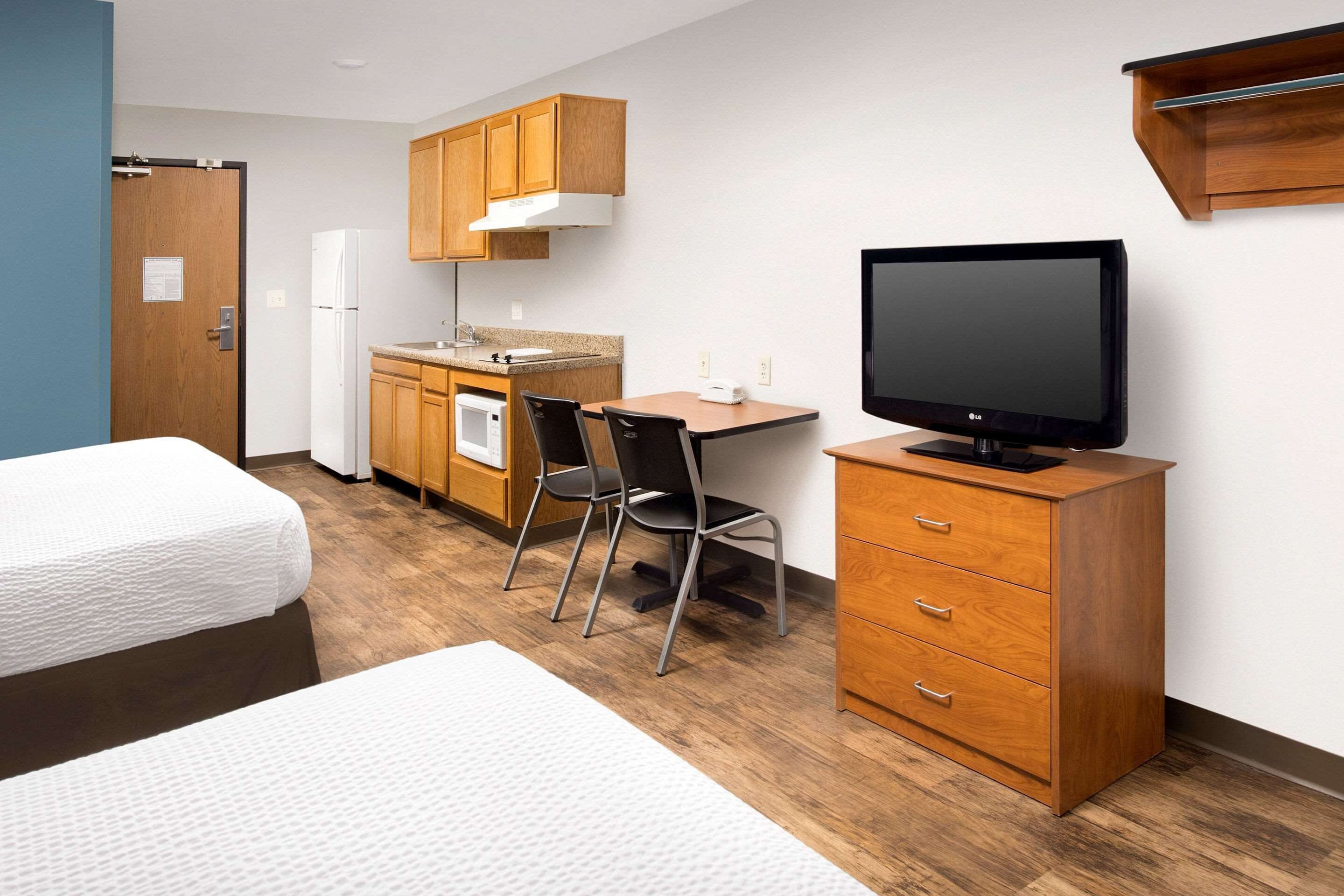 A compact hotel room with a kitchenette, a bed, a desk with a chair, and a TV on a cabinet.