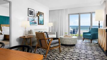 A modern hotel room with a large balcony, a round coffee table, a sofa, and a dining area.