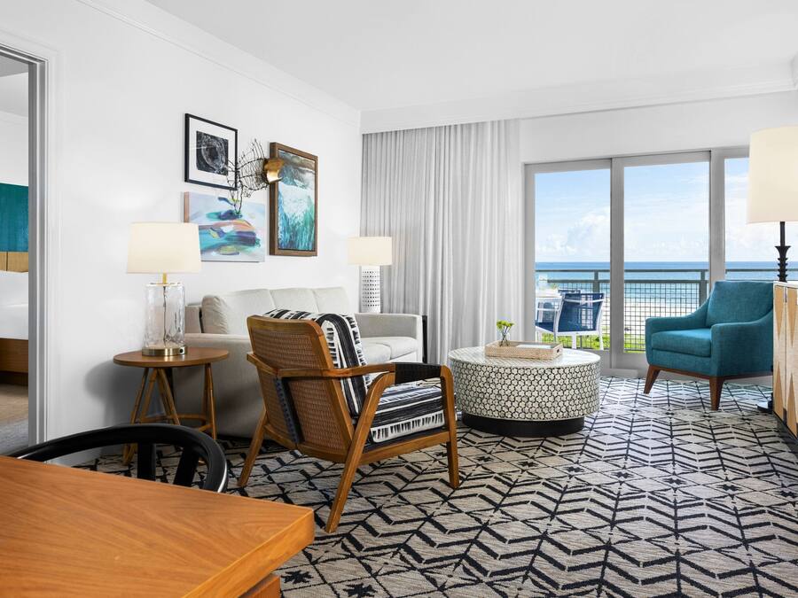 A modern hotel room with a large balcony, a round coffee table, a sofa, and a dining area.