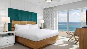 A modern hotel room with a large bed, a ceiling fan, a view of the sea, and a balcony with a chair.