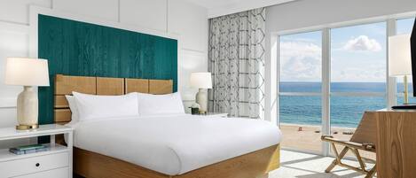 A modern hotel room with a large bed, a ceiling fan, a view of the sea, and a balcony with a chair.