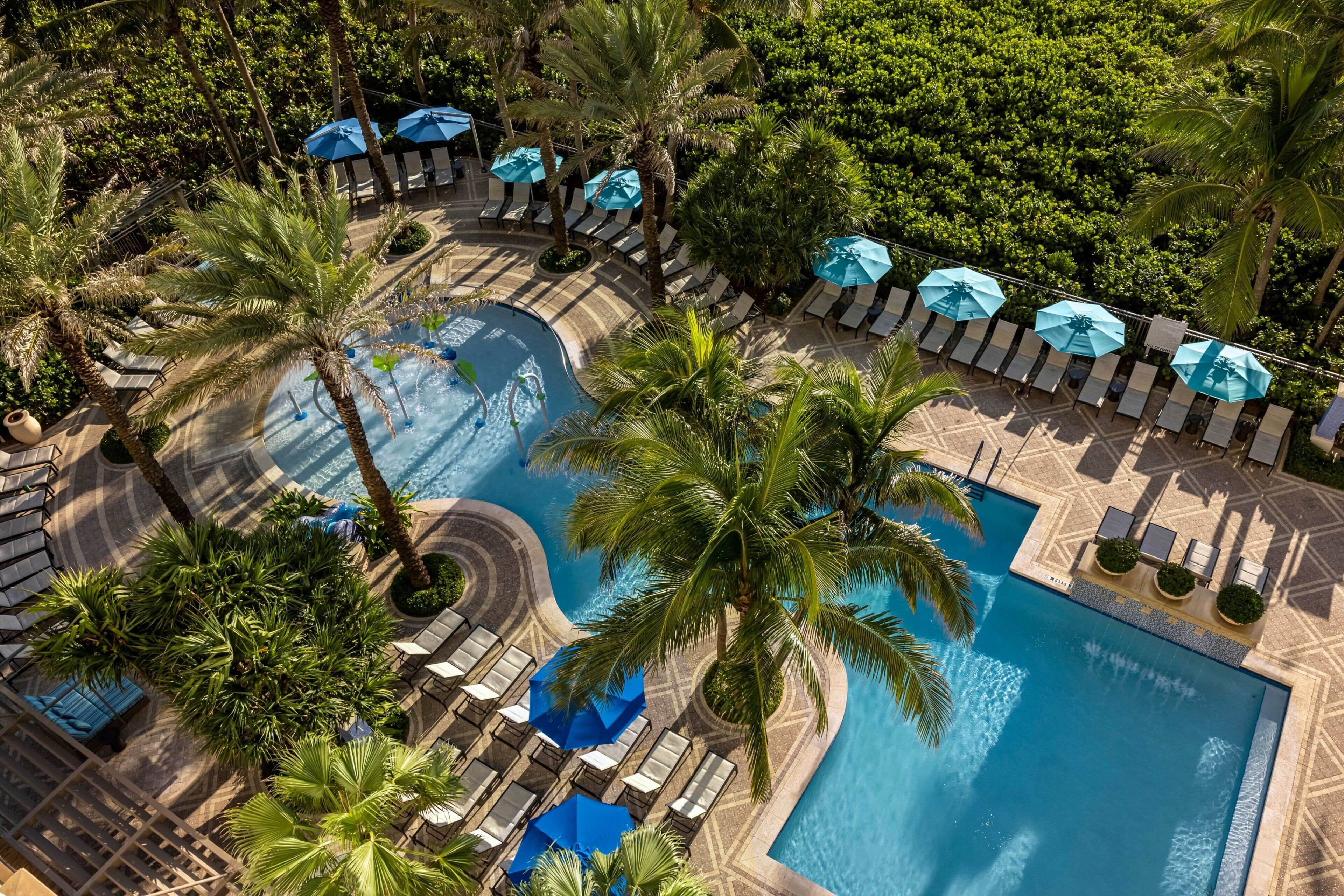 An aerial view of a resort with a large pool, sun loungers, and palm trees.