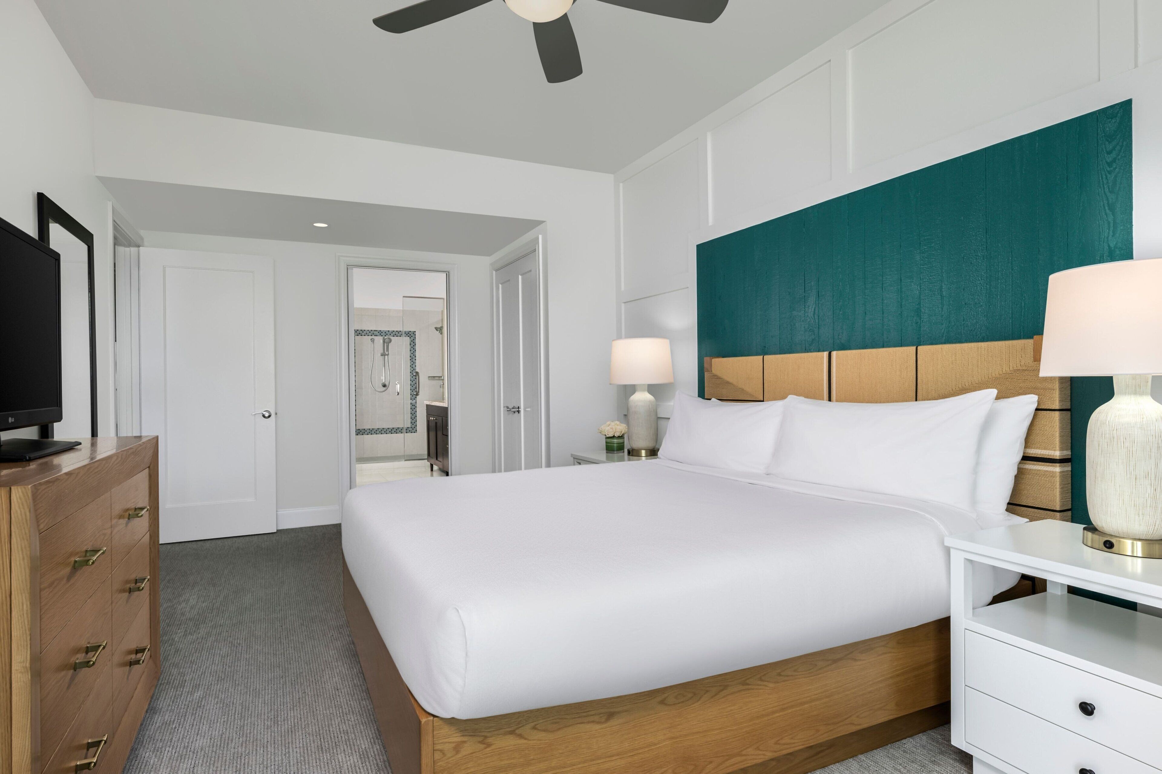 A hotel room with a large bed, wooden headboard, white bedding, a TV on a wooden dresser, and a ceiling fan.
