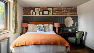 A bedroom with a brick wall, a bed with an orange blanket, two bedside lamps, a round mirror, and a desk with a chair.