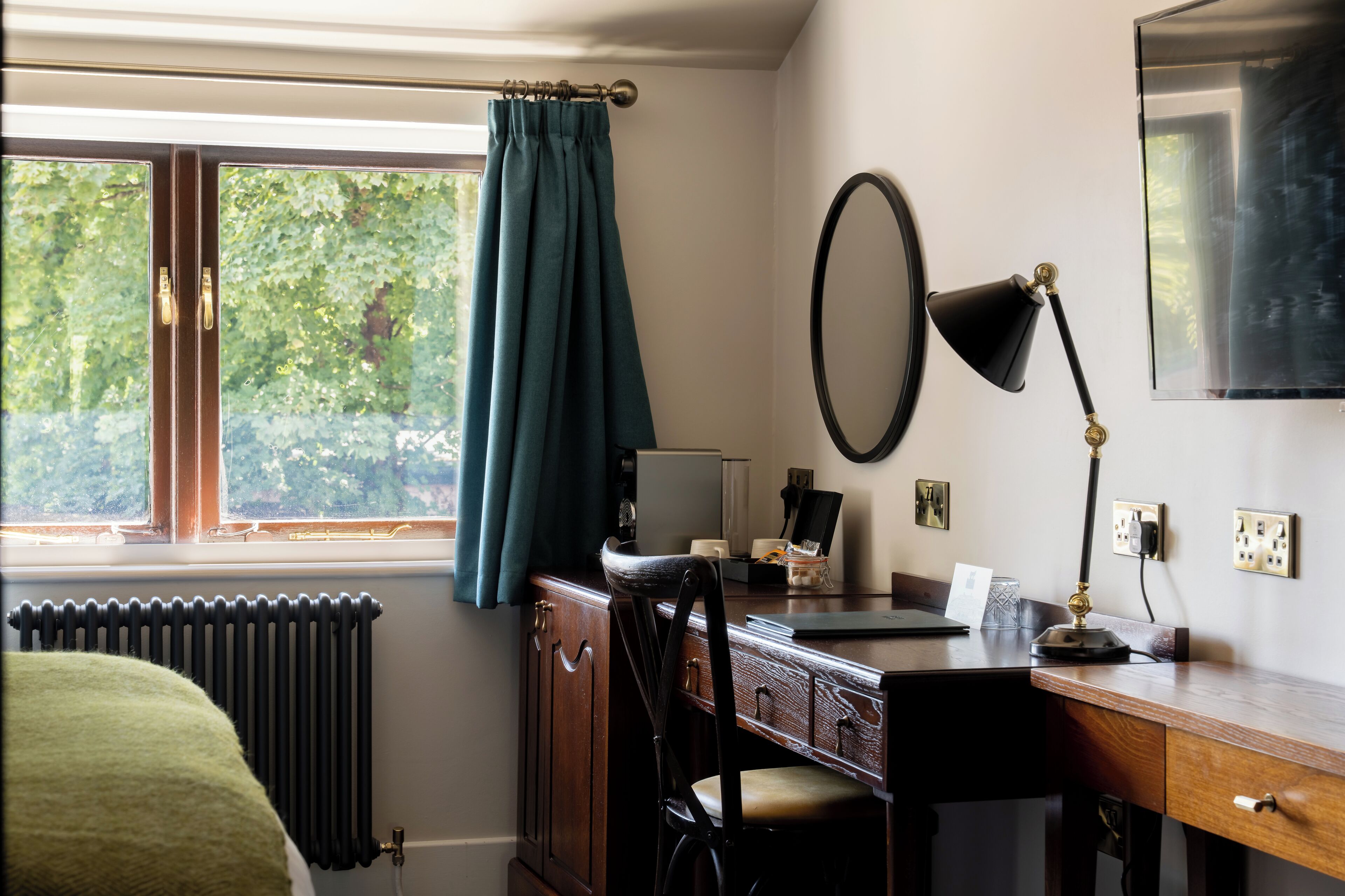 Una habitación con un escritorio de madera, una silla, un radiador, una ventana con cortinas, una lámpara de pared y un espejo.