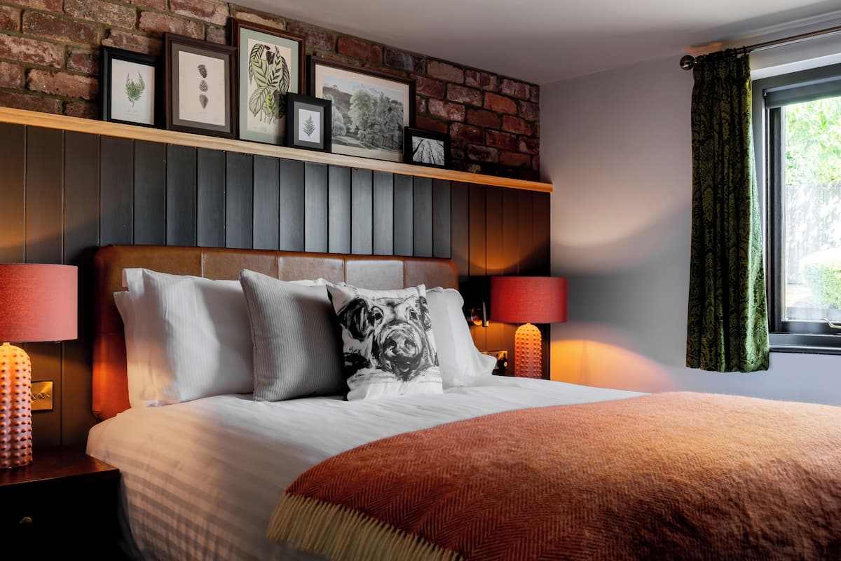 Una camera d'hotel con un muro in mattoni, un letto con biancheria bianca e una coperta rossa, due abat-jour e stampe botaniche incorniciate sopra il letto.