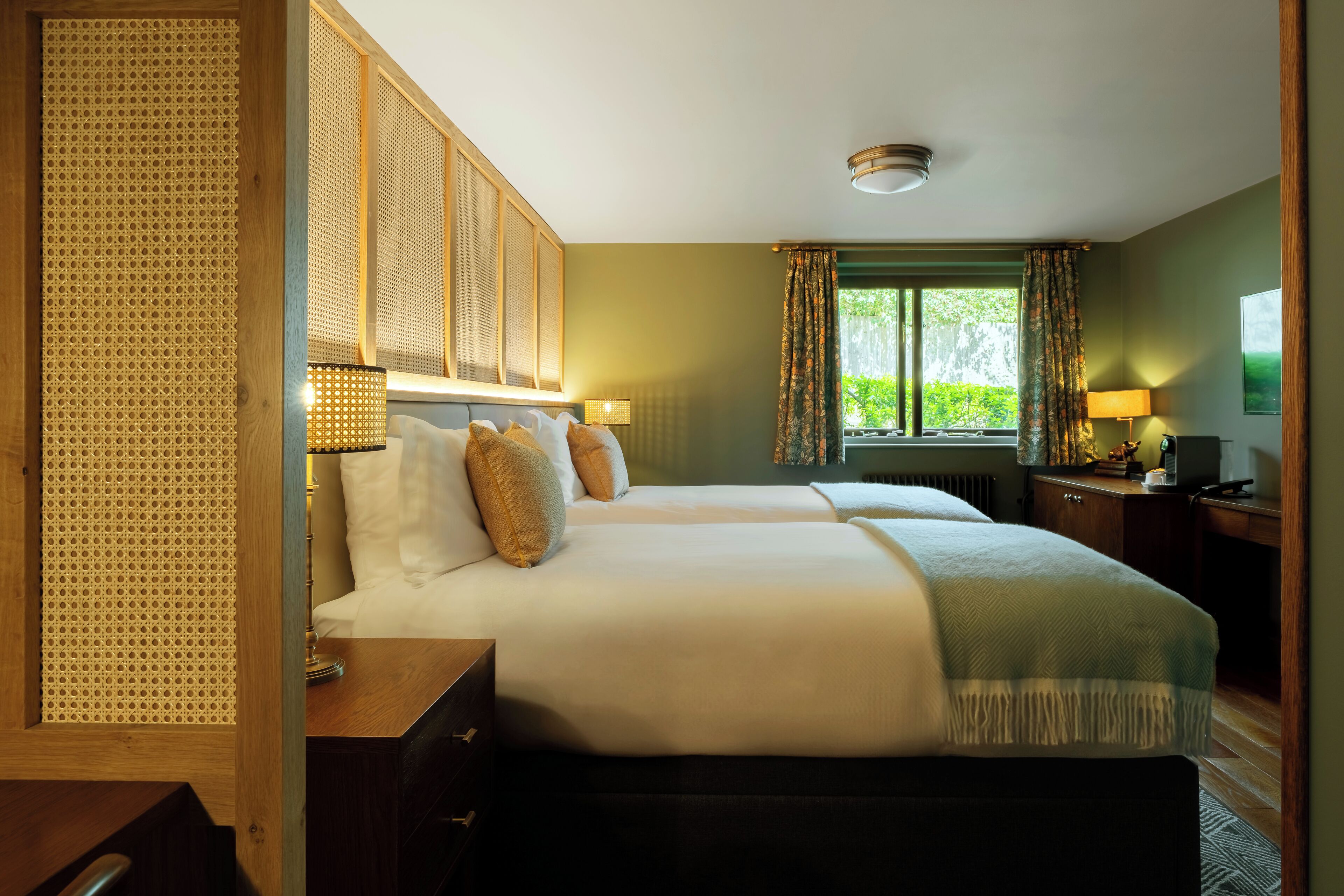 A hotel room with two beds, a wooden headboard, a desk, a chair, a lamp, and a window with curtains.