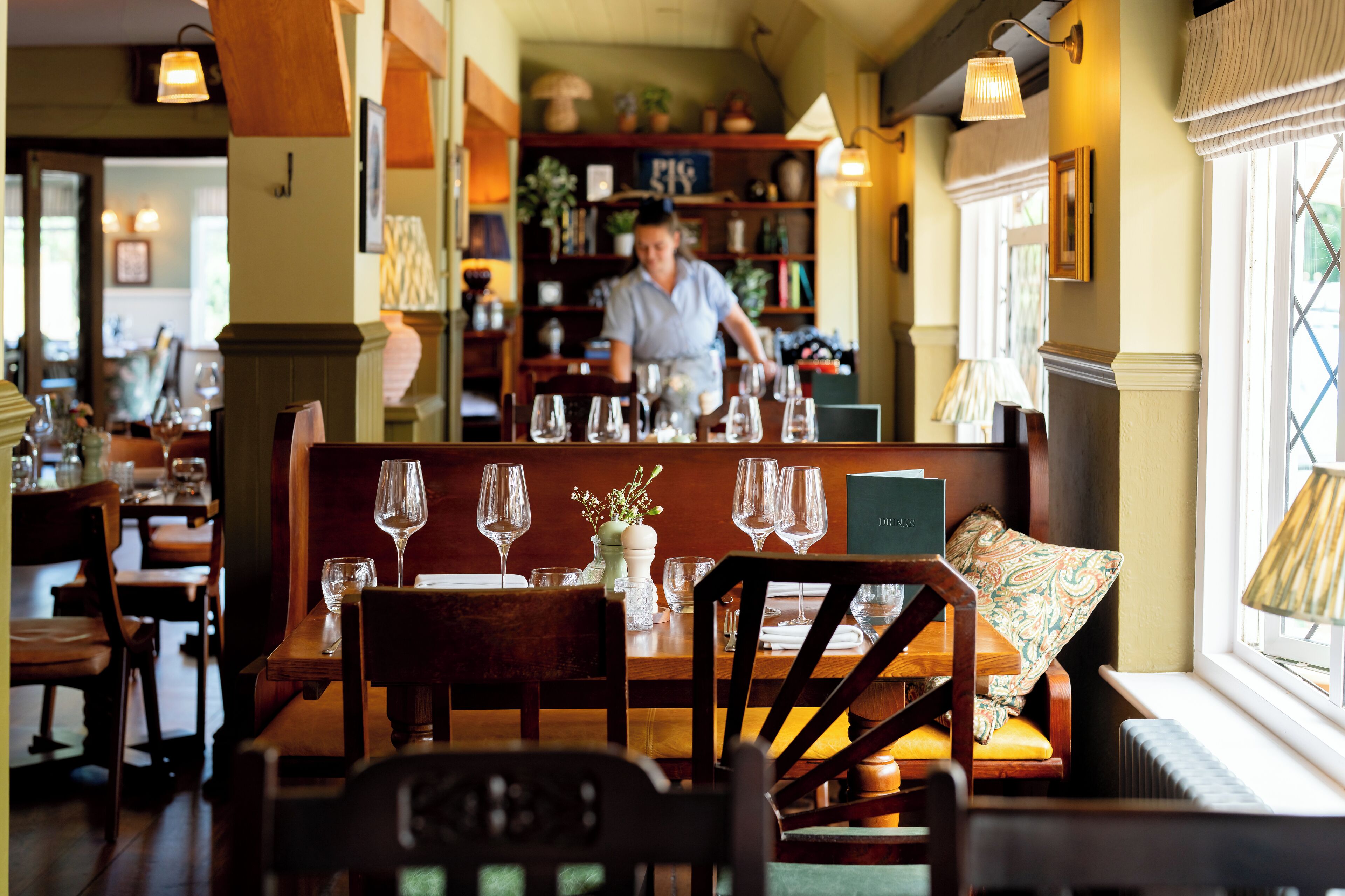 A restaurant interior with wooden furniture, a woman in the background, and a window with a lamp.