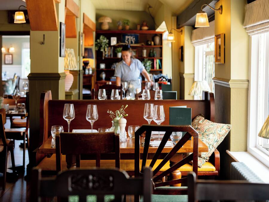Interior de restaurante con muebles de madera, una mujer al fondo y una ventana con lámpara.