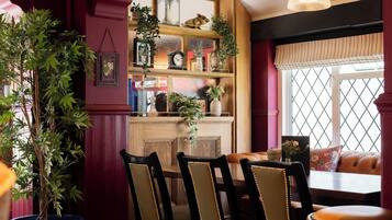A cozy dining area with wooden chairs, a table, and a bench. There is a bookshelf with various items and a potted plant.