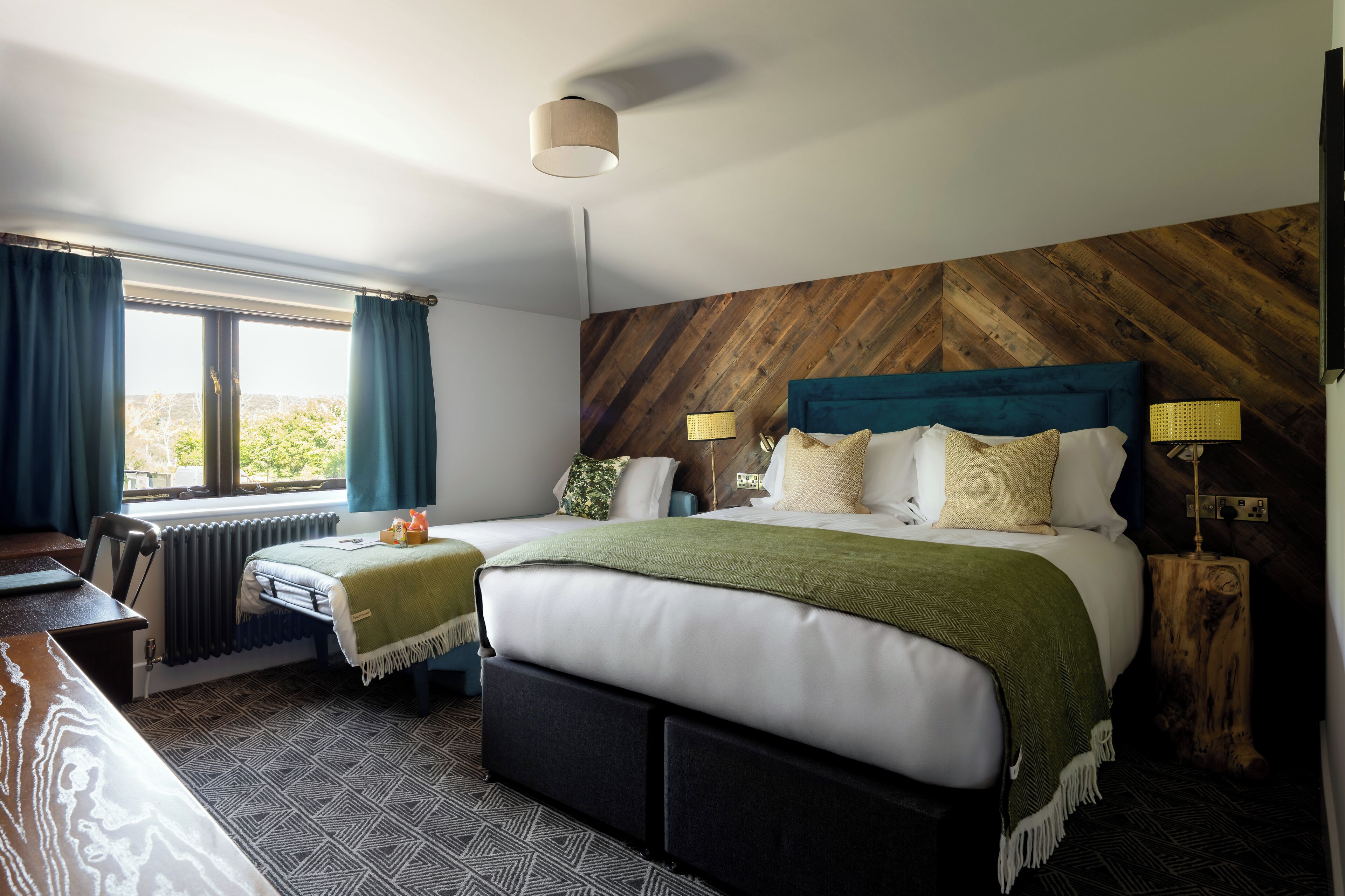 A bedroom with a bed, a desk, a chair, a window with curtains, a radiator, and a wooden wall.