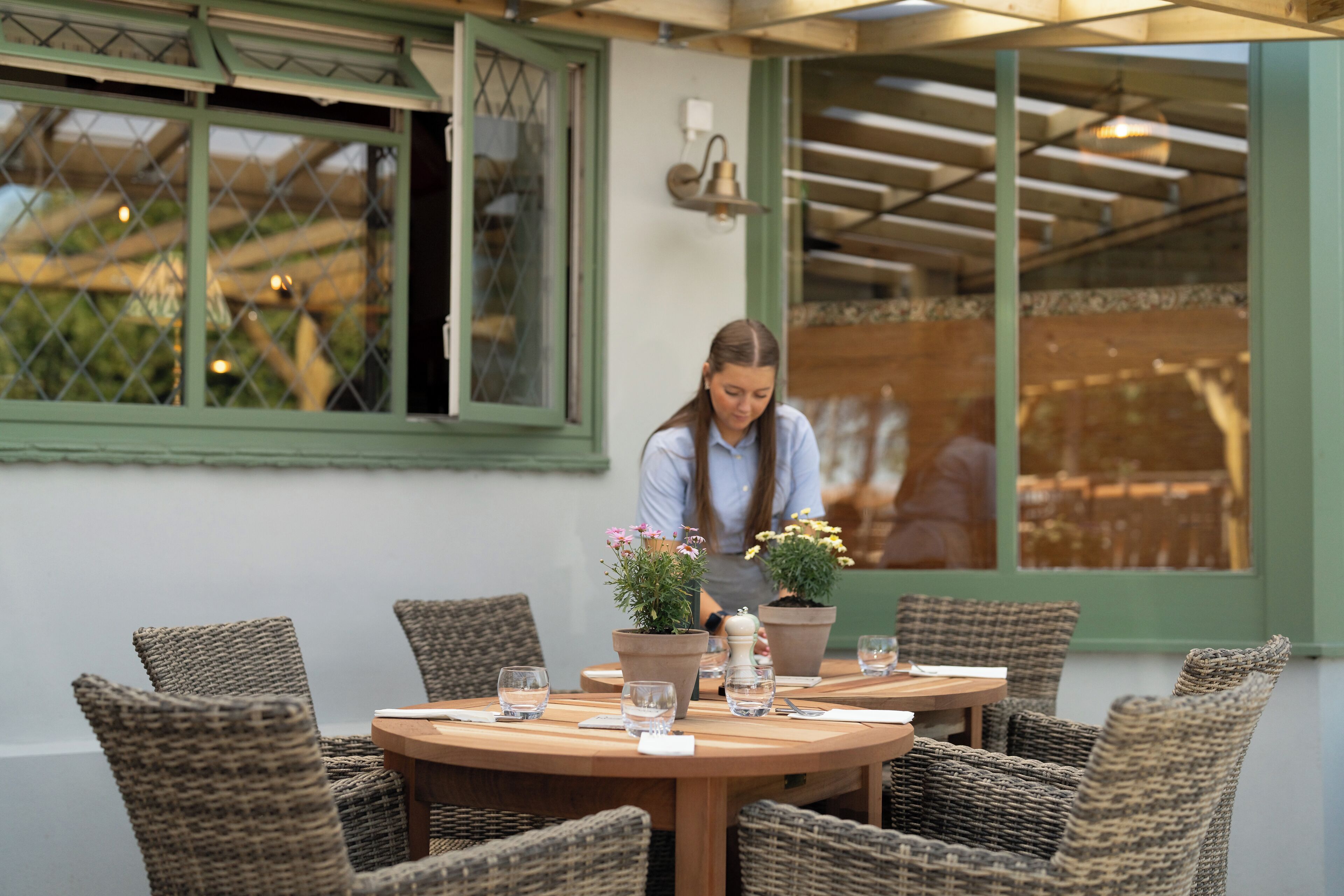 A round wooden table with wicker chairs, potted plants, and a person arranging items.
