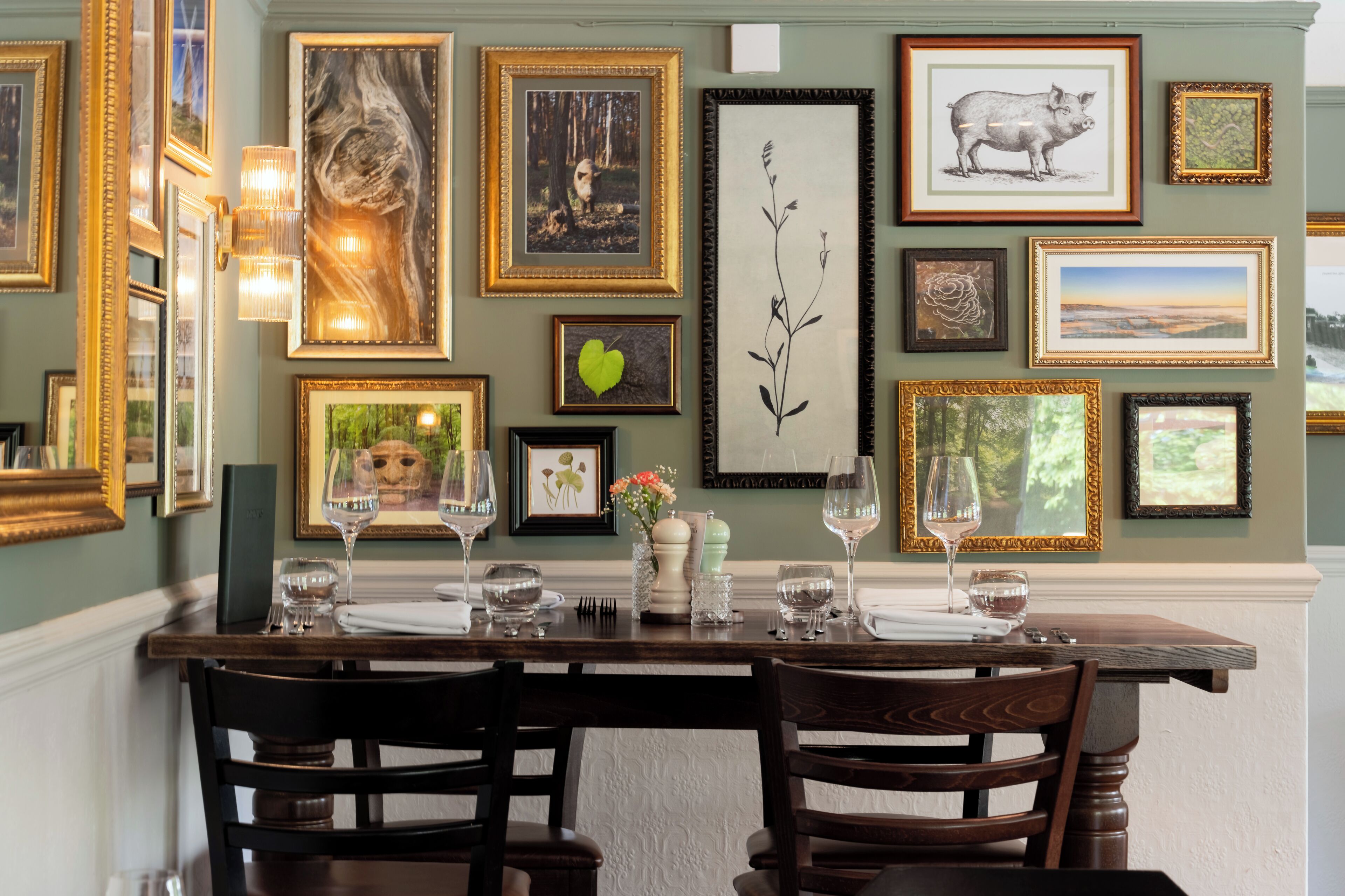 A dining area with a wooden table set for two, surrounded by framed artwork on the walls.