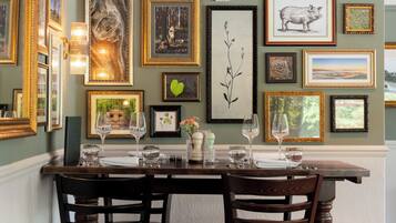 A dining area with a wooden table set for two, surrounded by framed artwork on the walls.