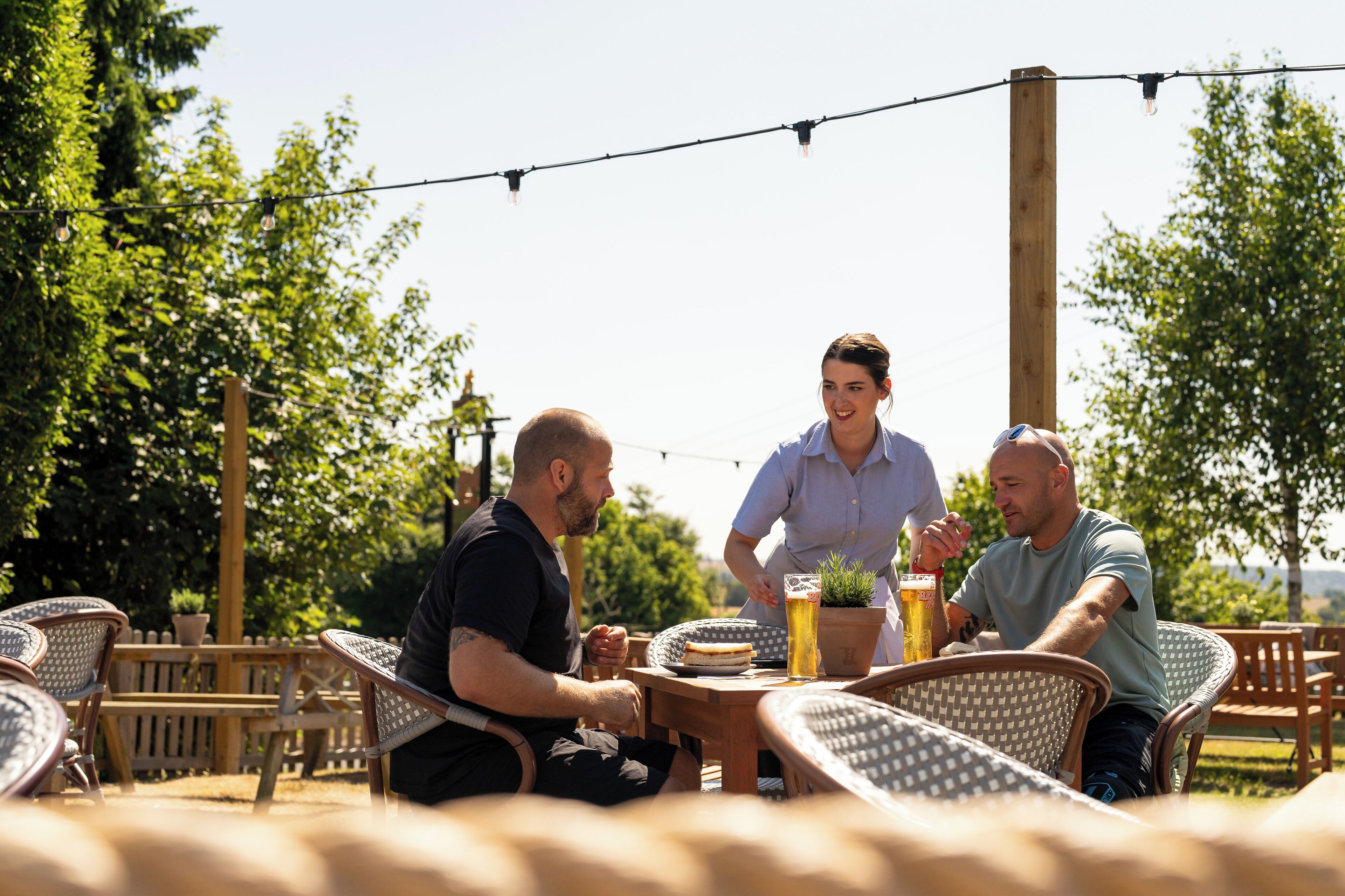 A group of people seated at an outdoor table with wicker chairs, a wooden table, and a potted plant.
