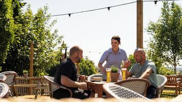 A group of people seated at an outdoor table with wicker chairs, a wooden table, and a potted plant.