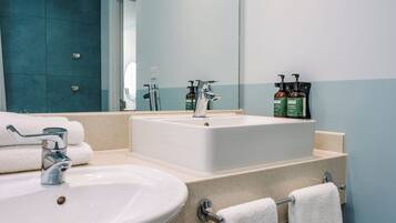 Un baño con un lavamanos blanco, un espejo grande y una encimera color beige. Hay dos botellas de champú y una botella de loción sobre la encimera.