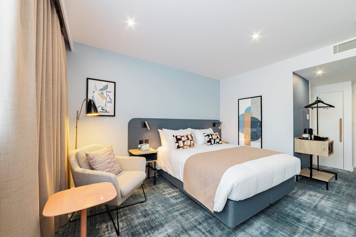 A modern hotel room with a large bed, a chair, a desk, and a wardrobe.