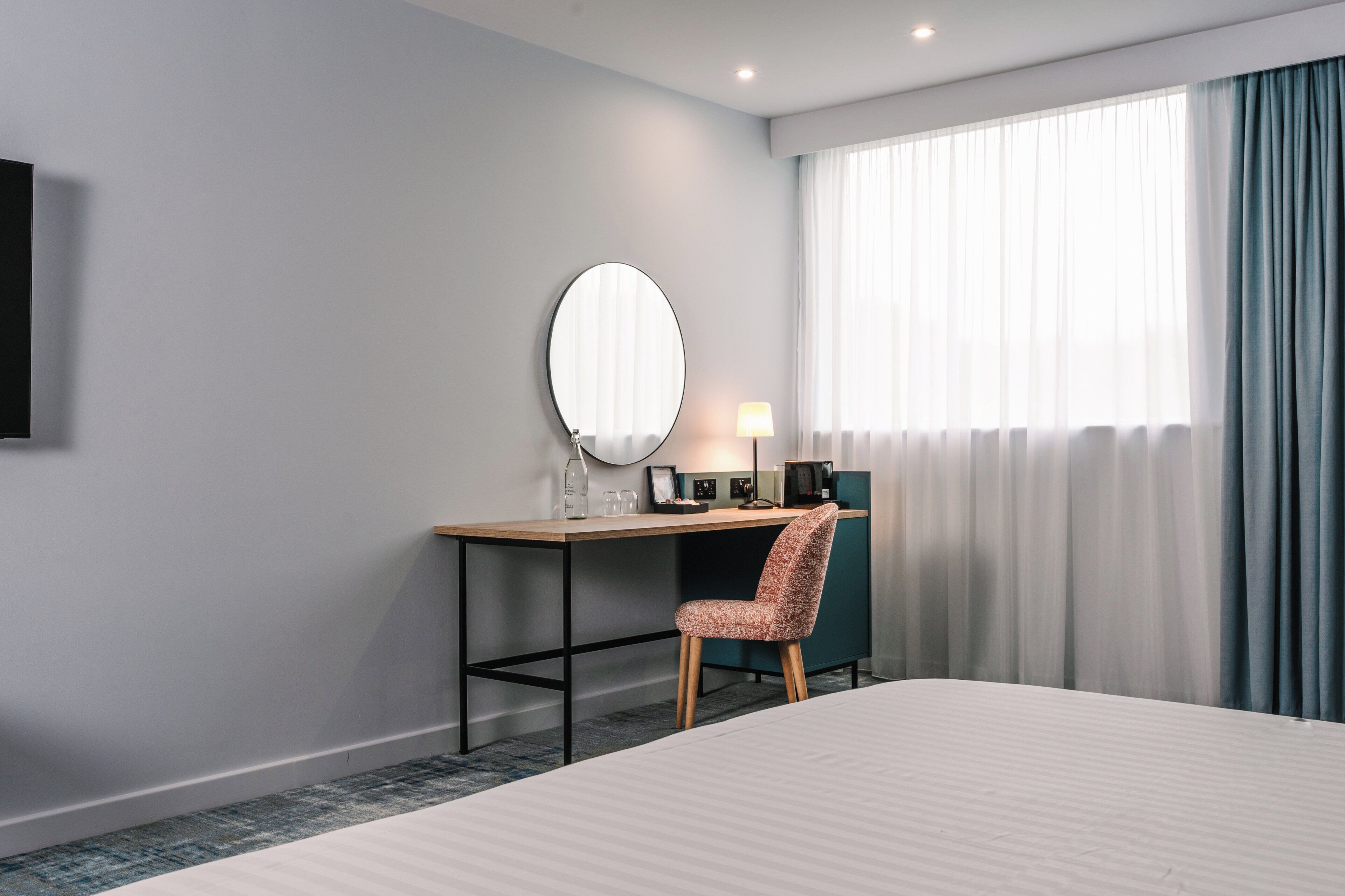 Een hotelkamer met een bed, een bureau met stoel, een ronde spiegel, een lamp en een raam met gordijnen.