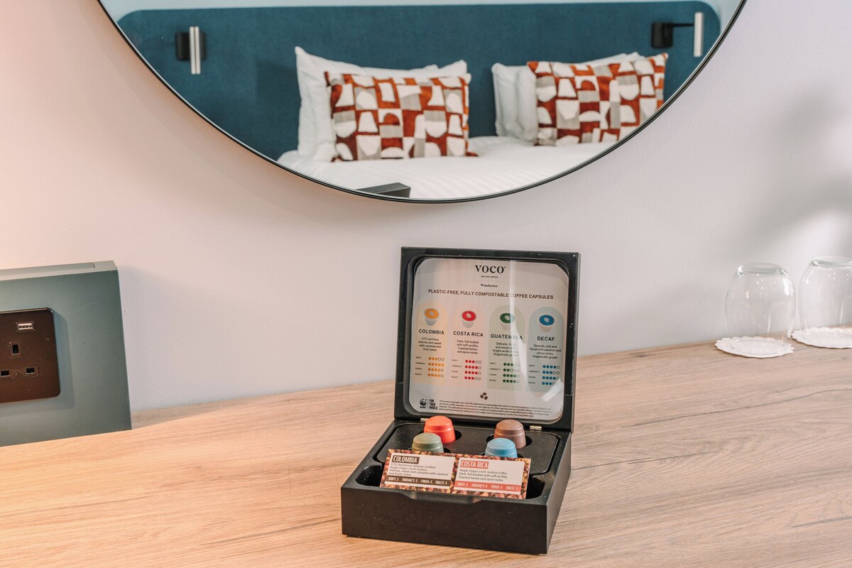 Une chambre d’hôtel avec une table de chevet en bois, un miroir reflétant un lit agrémenté d’oreillers à motifs et un présentoir à capsules de café.