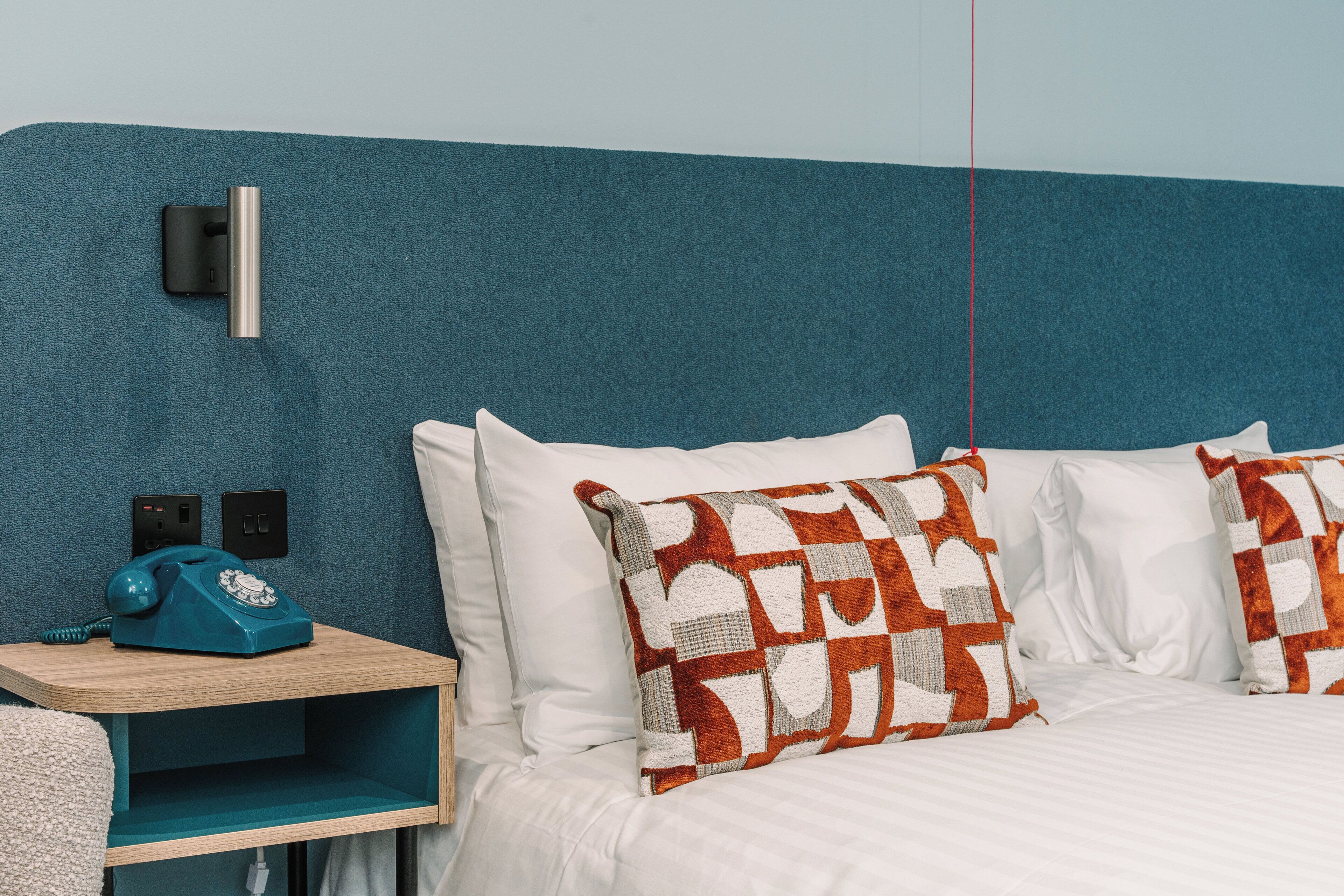 Une chambre d’hôtel avec un mur d’accent de couleur turquoise, une table de chevet en bois surmontée d’un téléphone bleu d’époque, du linge de lit blanc et des coussins décoratifs à motifs.