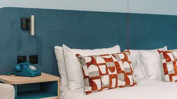 Une chambre d’hôtel avec un mur d’accent de couleur turquoise, une table de chevet en bois surmontée d’un téléphone bleu d’époque, du linge de lit blanc et des coussins décoratifs à motifs.