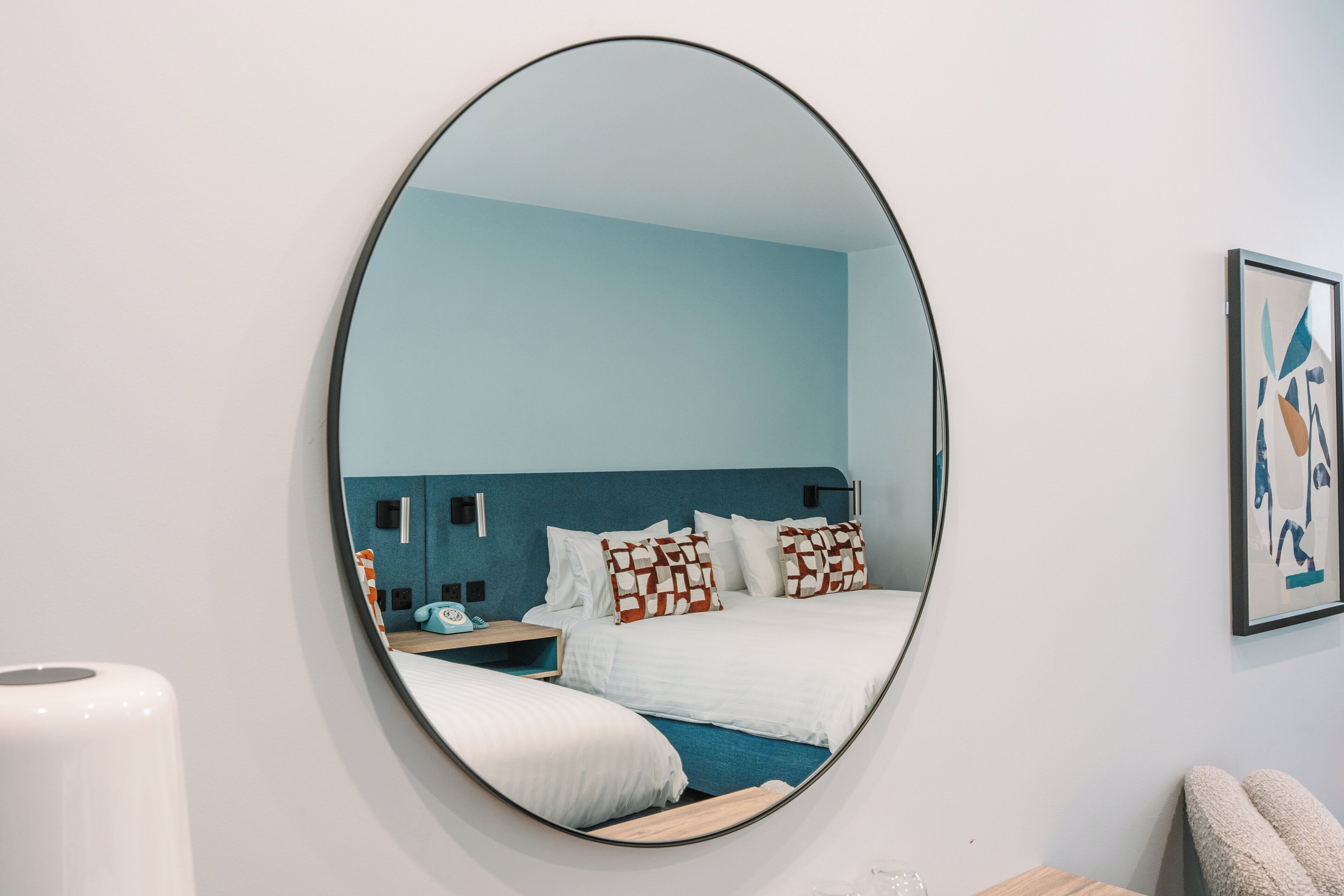 A hotel room with a large round mirror, a bed with patterned pillows, a bedside table with a lamp, and a framed abstract artwork on the wall.