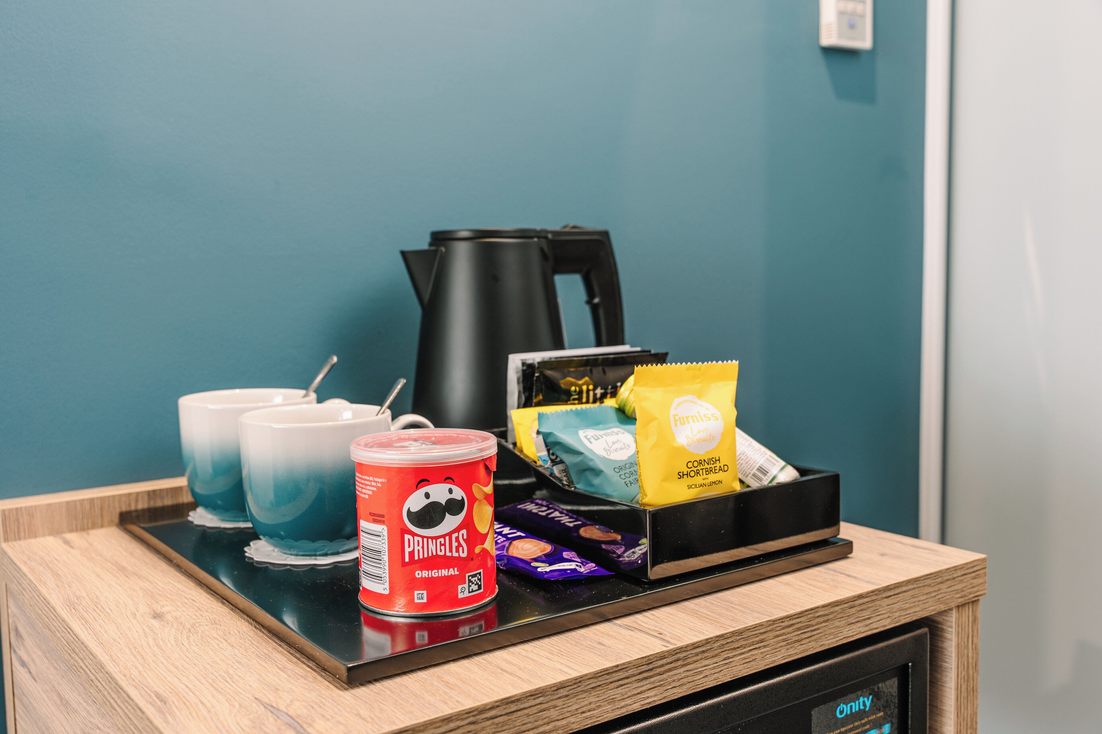 Una bandeja con cafetera, dos tazas, un bote de Pringles, una bolsa de galletas de maíz y una caja de bombones.