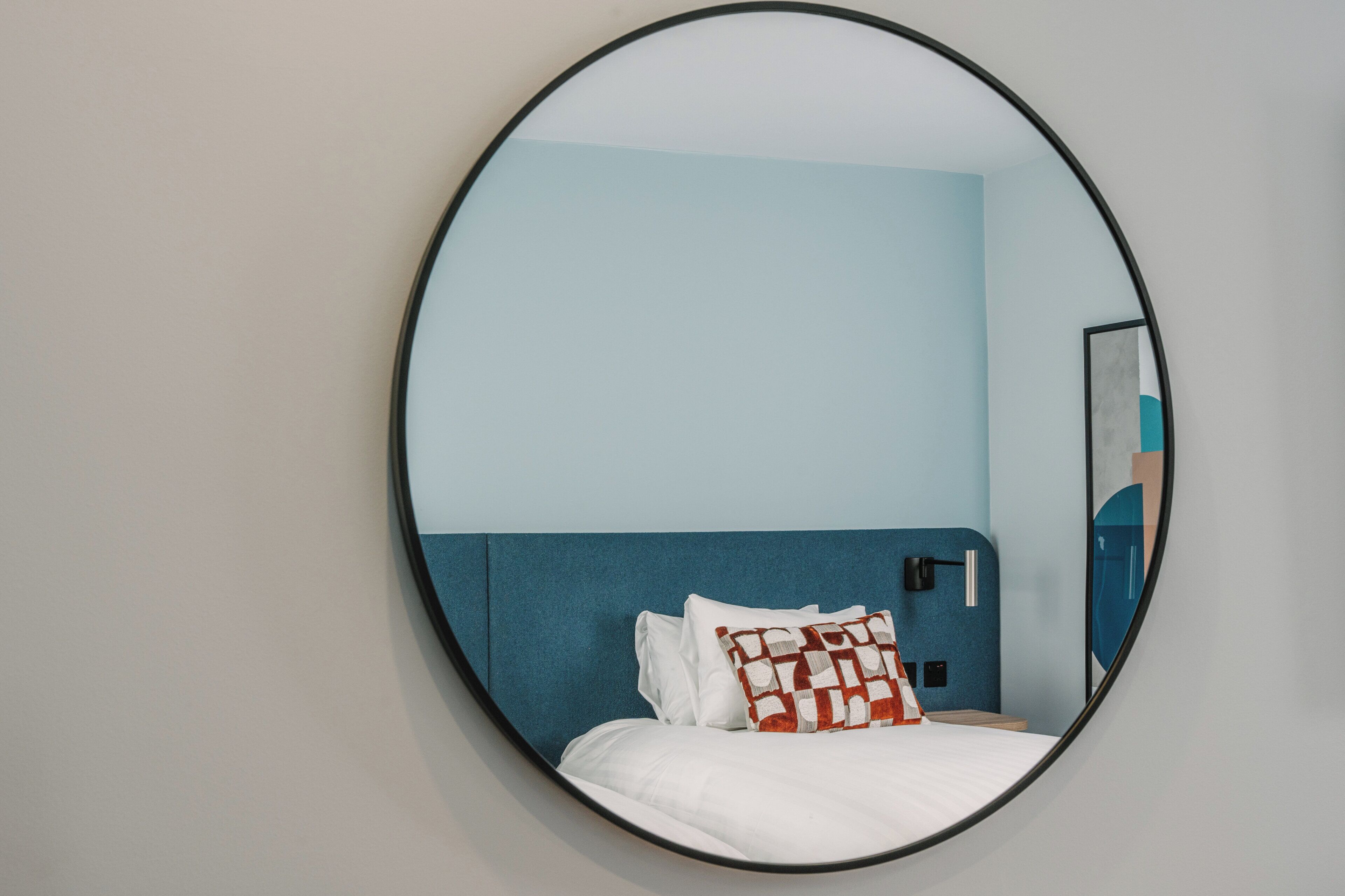 Un miroir rond reflète un lit doté d’une tête de lit bleue, de draps blancs et d’un coussin à motifs rouges et blancs.