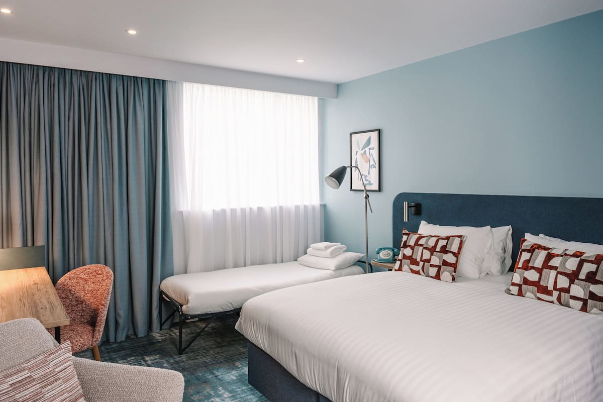 A hotel room with a large bed, two smaller beds, a desk, a chair, and a window with curtains.