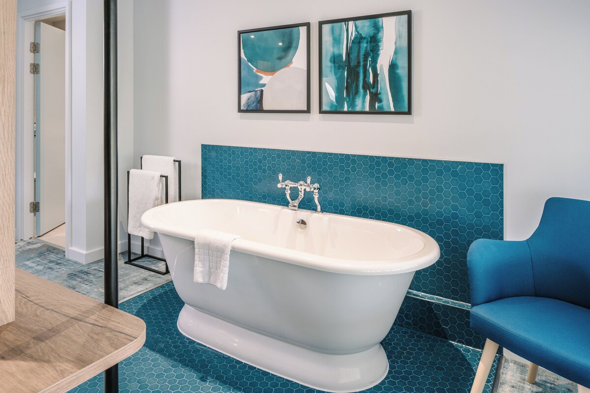 Une salle de bain moderne avec une baignoire sur pieds, une chaise bleue et deux tableaux abstraits accrochés au mur.