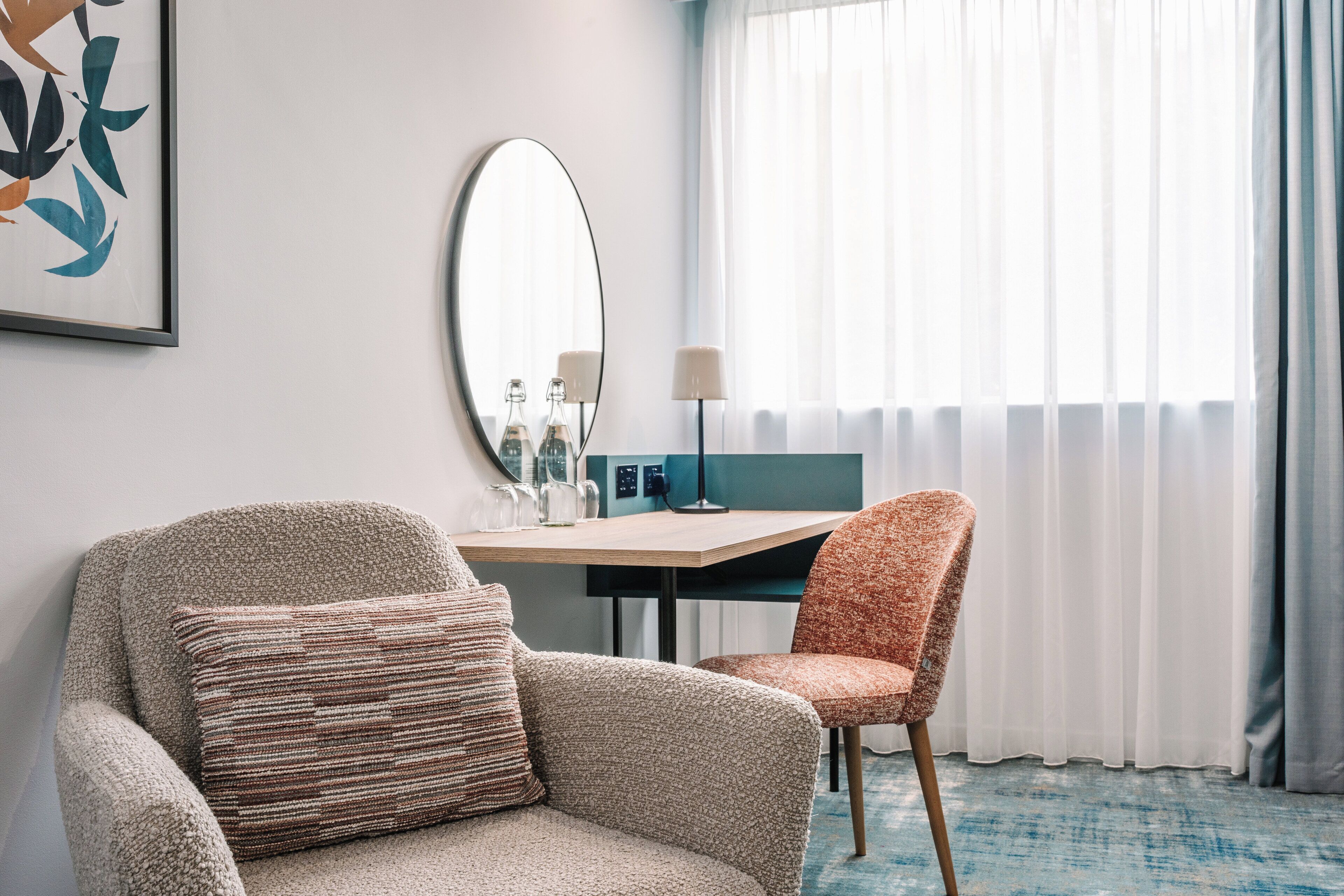 A hotel room with a sofa, a chair, a desk, a mirror, and a window with curtains.