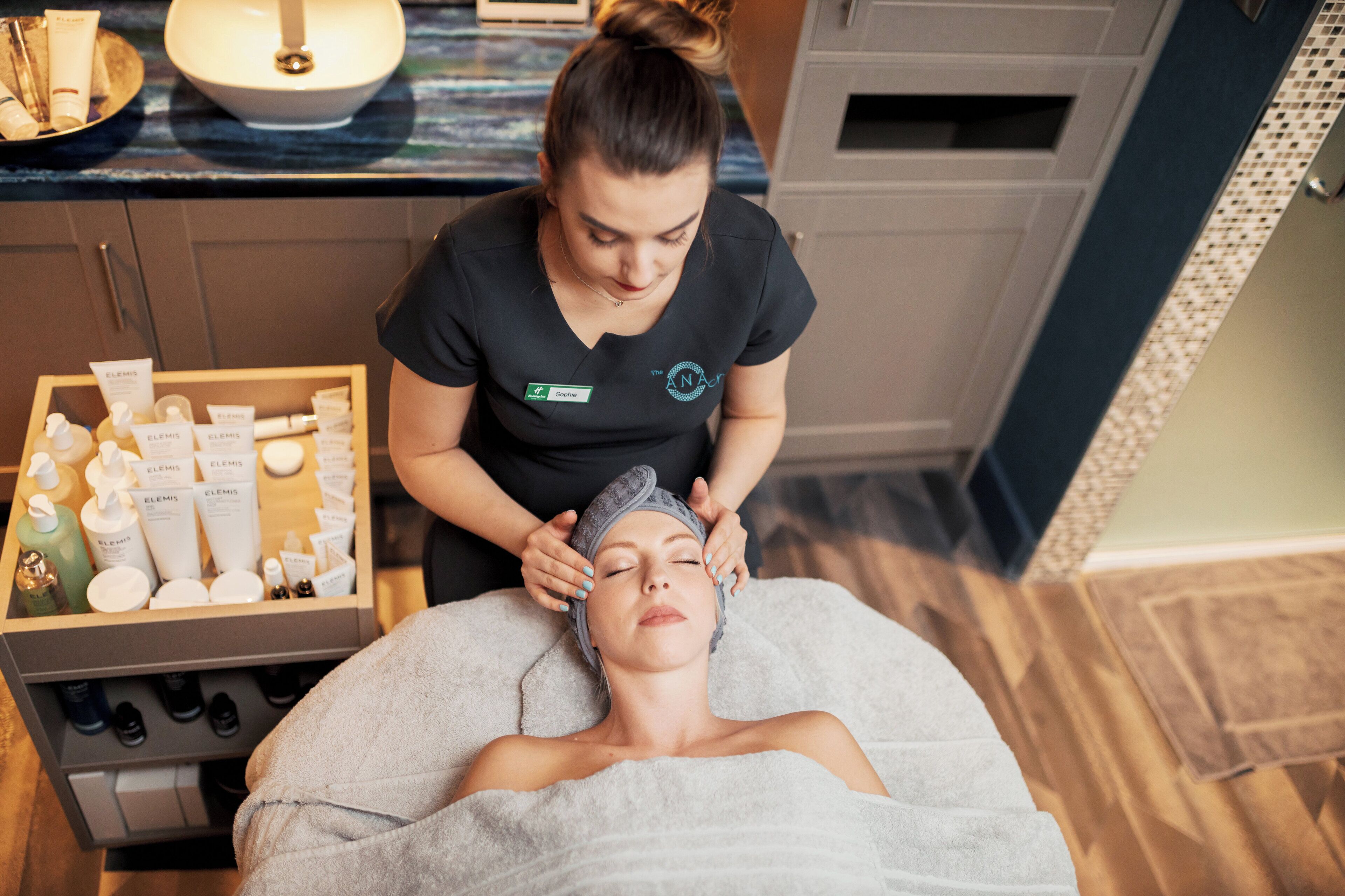 A spa setting with a massage table, a woman performing a head massage, and a shelf with various skincare products.