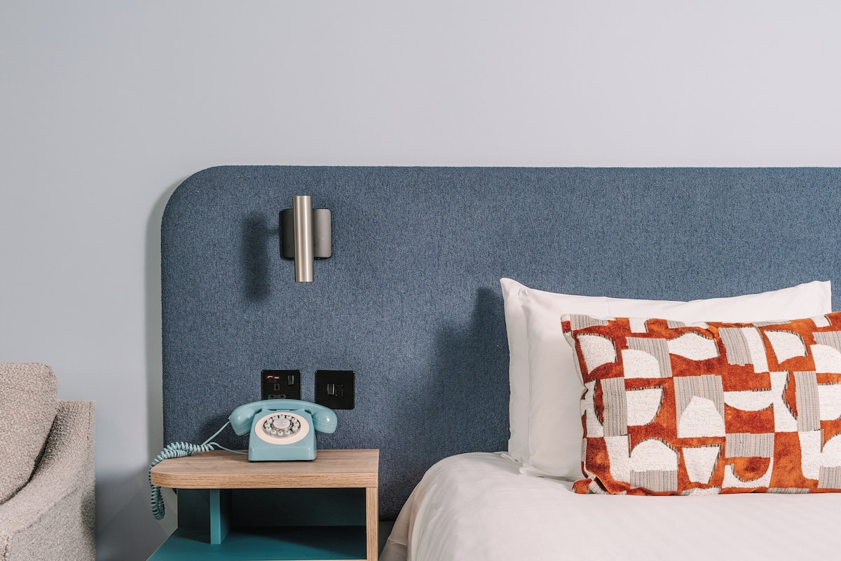 A hotel room with a blue textured headboard, a wooden nightstand with a vintage-style telephone, a white pillow with a geometric pattern, and a beige armchair.